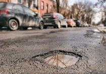 An up-close view of a small, rain-filled pothole on a street. In the blurred background is a line of cars parked on a curb. 