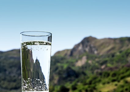 A clear glass of water with blue sky and green mountain landscape background 