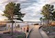 Rendering shows people walking along a wooden boardwalk. To the left is a body of water, and there are yachts in the water. To the right of the boardwalk are trees and a pavilion.