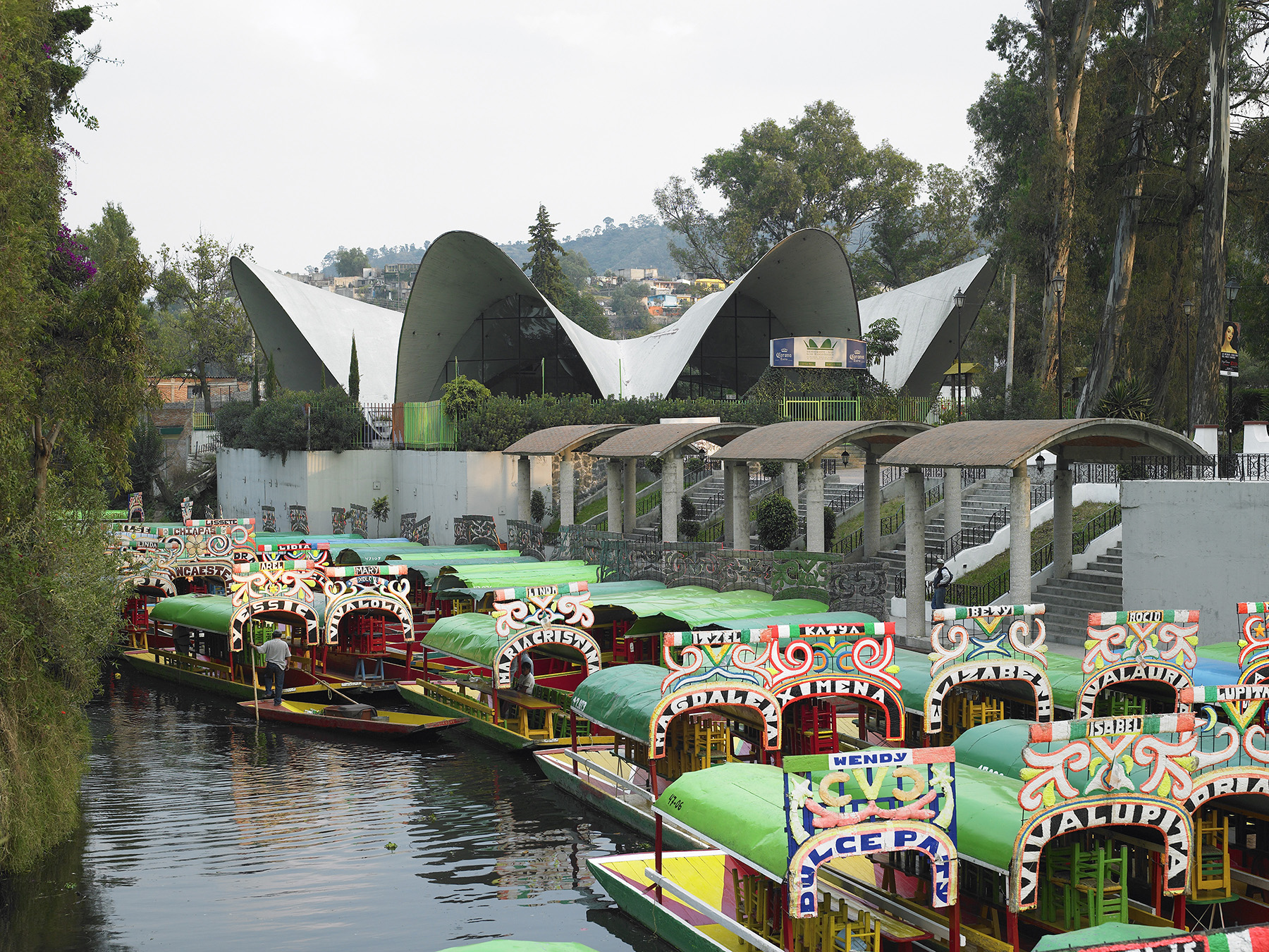 Bright-colored boats float in the water in the foreground. In the background is a circular structure topped by a wavy roof. 