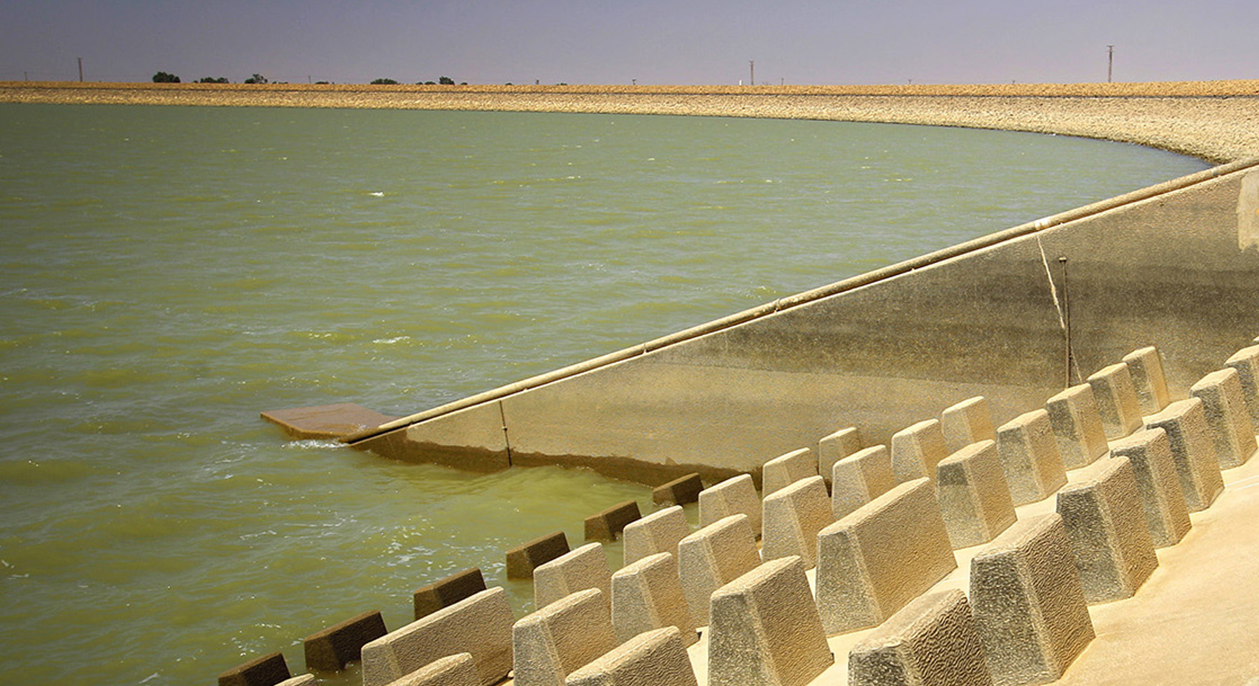 Photograph shows a large body of water surrounded by concrete. Blocks of concrete have been inserted into a concrete base. 