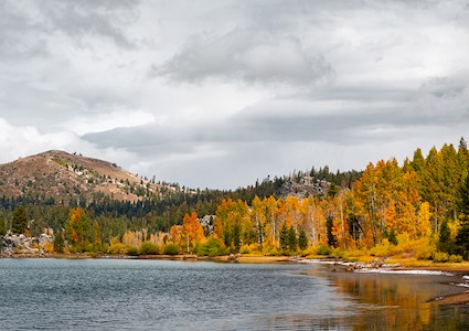 Photo shows a lake and in the background are autumnal-colored tress and a small mountain. 