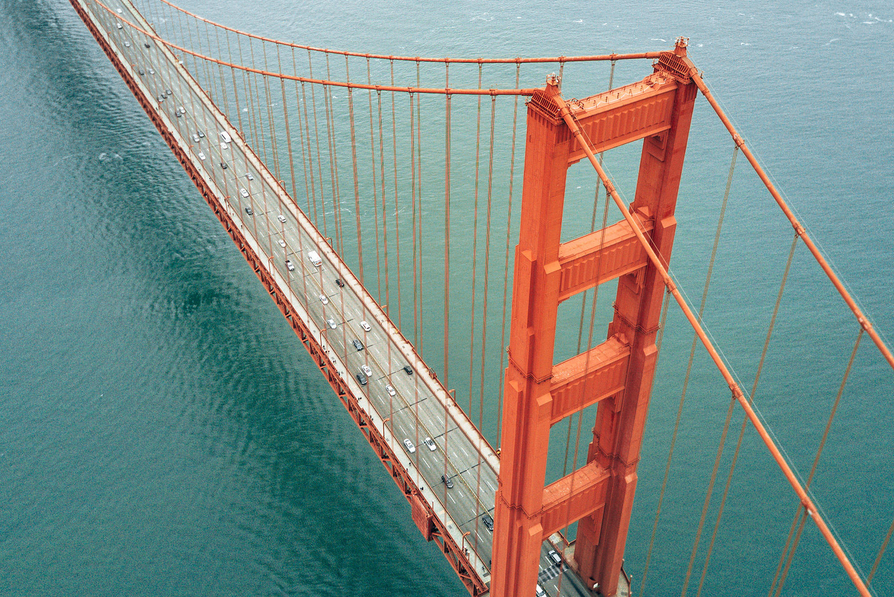 Red-colored tower of a cable-stayed bridge is shown. The bridge spans a large body of water. Cars travel on the bridge. 