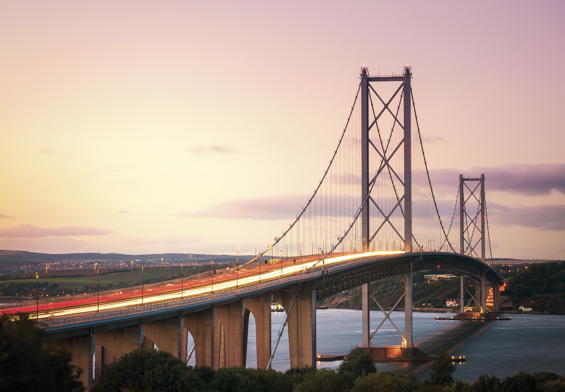 A cable-stayed bridge is shown at night. The roadway appears to be lit. Water flows beneath the structure. 