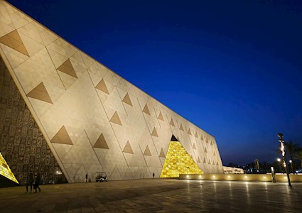 A long and tall stone wall has stone triangles embedded in it. in the center of the wall is a triangular shaped structure that is lit from within. 