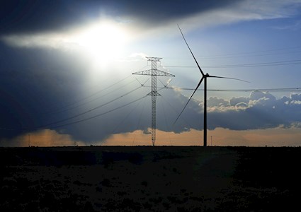 A power line structure and a wind mill are set in a desolate, night-time landscape. 