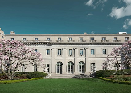 The stone facade of a multistory mansion is shown. There are flowers on the trees and steps leading down to beautiful green grass. 