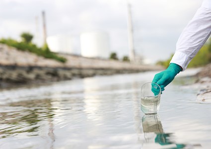 A green gloved hand for someone wearing a white protective suit takes a water sample using a clear beaker. 