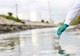 A green gloved hand for someone wearing a white protective suit takes a water sample using a clear beaker. 