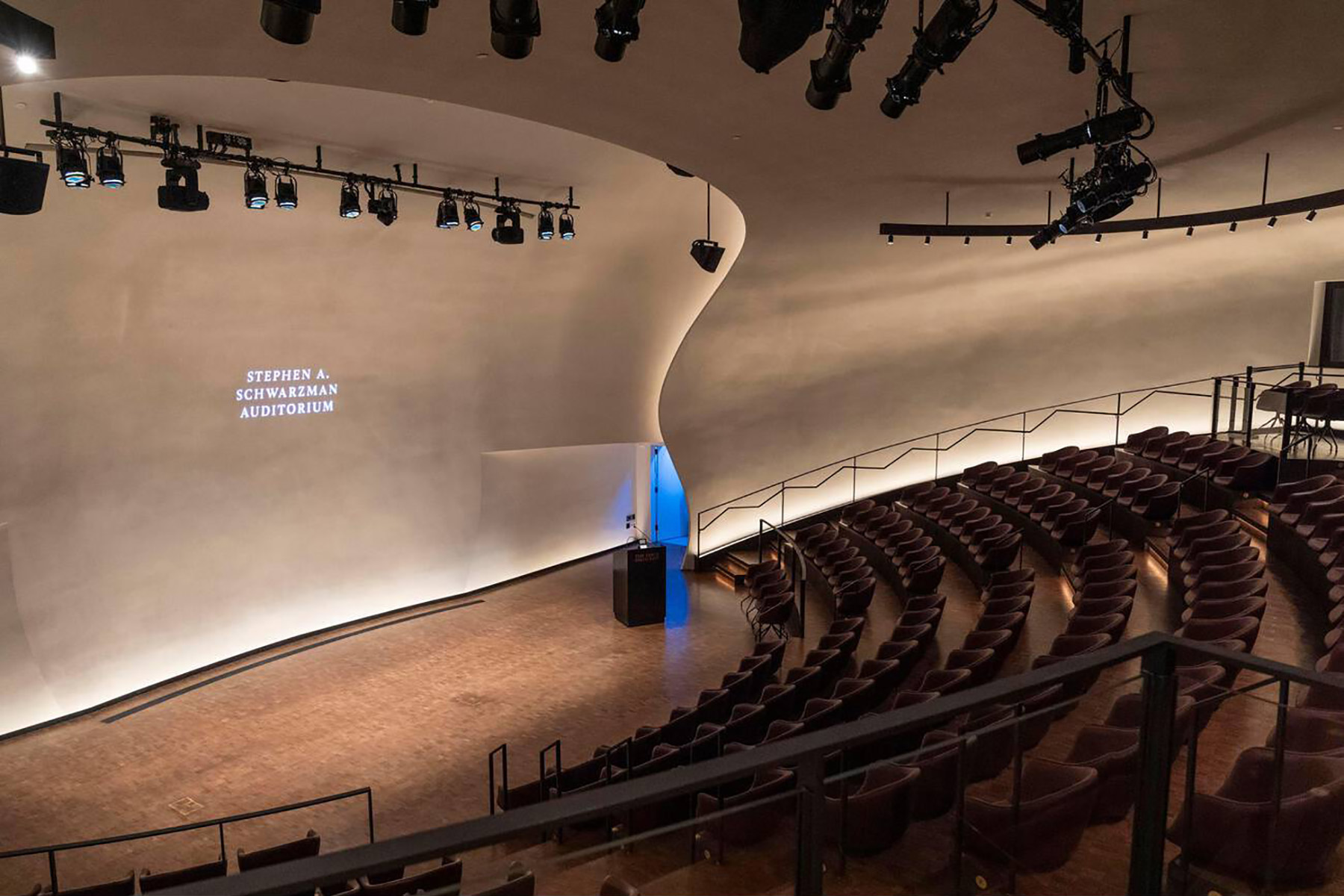 The room shown is dimly lit. There is a podium on the stage and stacked seating. 