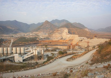 The photograph shows the massive dam under construction in an arid landscape. 