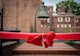 A blurred image of a six-ton bronze bell that hangs in a park, surrounded by brick buildings. A red bow is added over the image. 