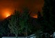 a house with green trees sits in the foreground while a wildfire rages in the distance in the background