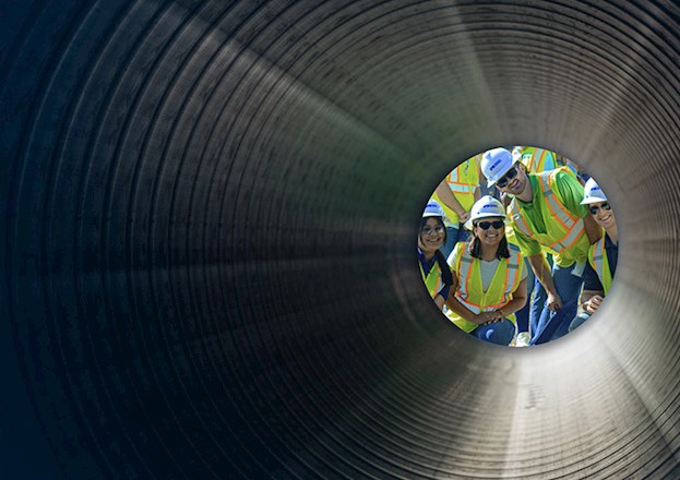 Four students wearing yellow safety vests, black sunglasses, and white hard hats smile into a ridged tunnel.