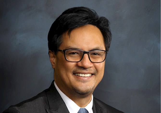 Studio headshot of an adult professional wearing glasses, a dark suit jacket, and a light-colored shirt, set against a dark textured background.